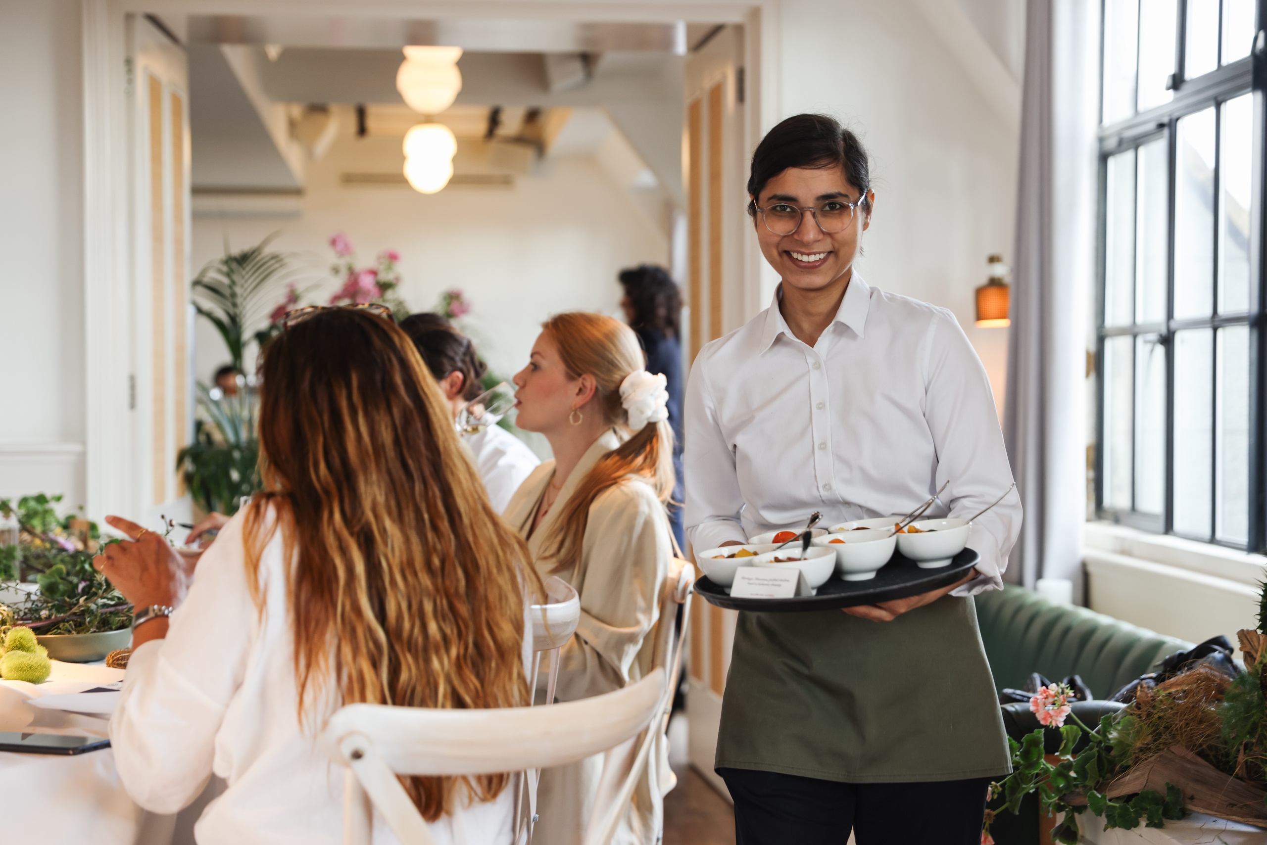 A smiling Maslow’s team member serves guests in a bright, plant-filled dining room, embodying the brand’s signature intuitive service and calm hospitality style.