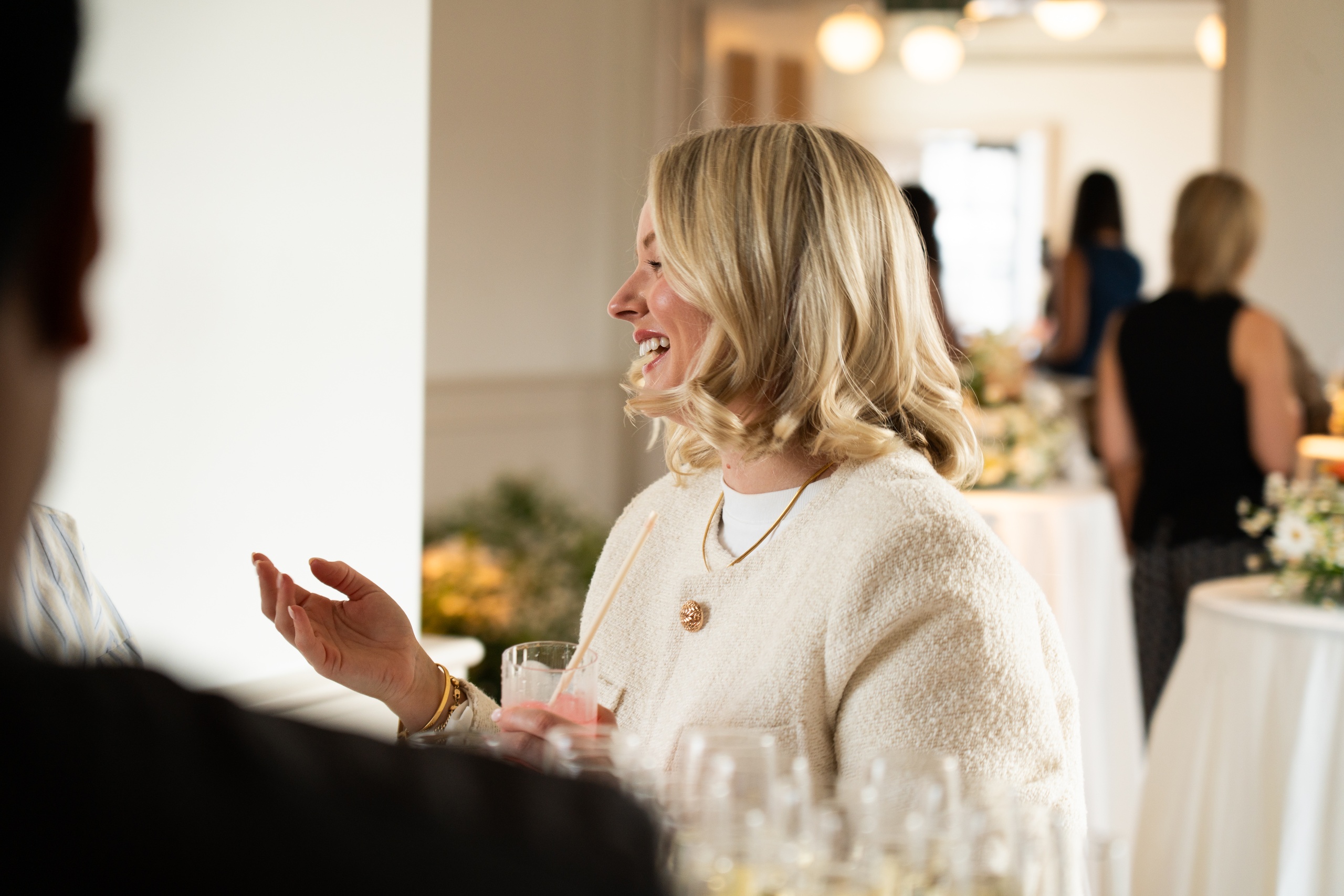 Smiling woman holding a cocktail and chatting during a private event in London, with elegant floral décor in the background.