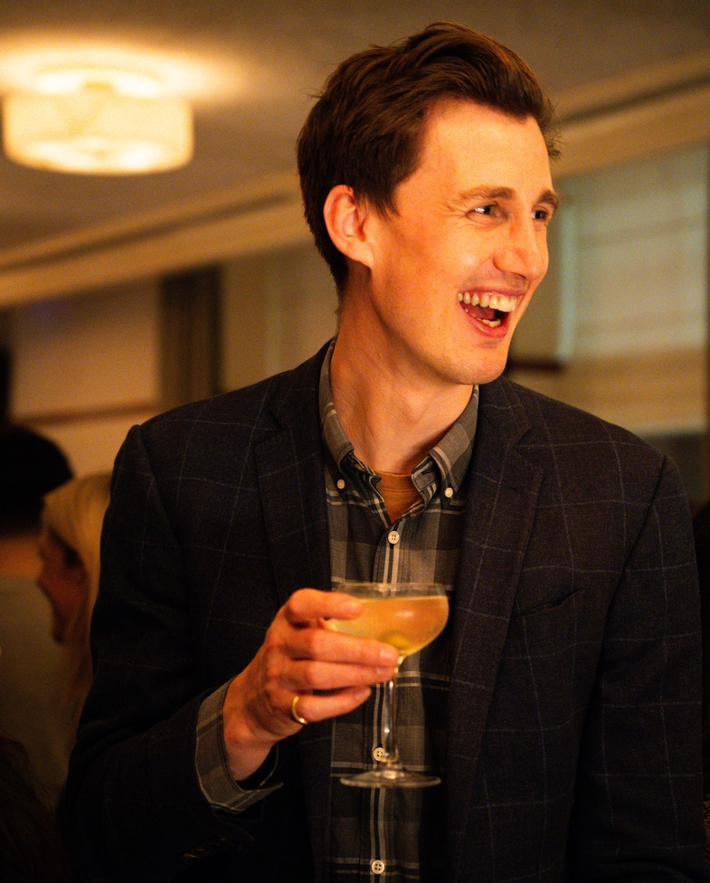 Smiling man in a checked blazer holding a cocktail at a Maslow’s private event in London.
