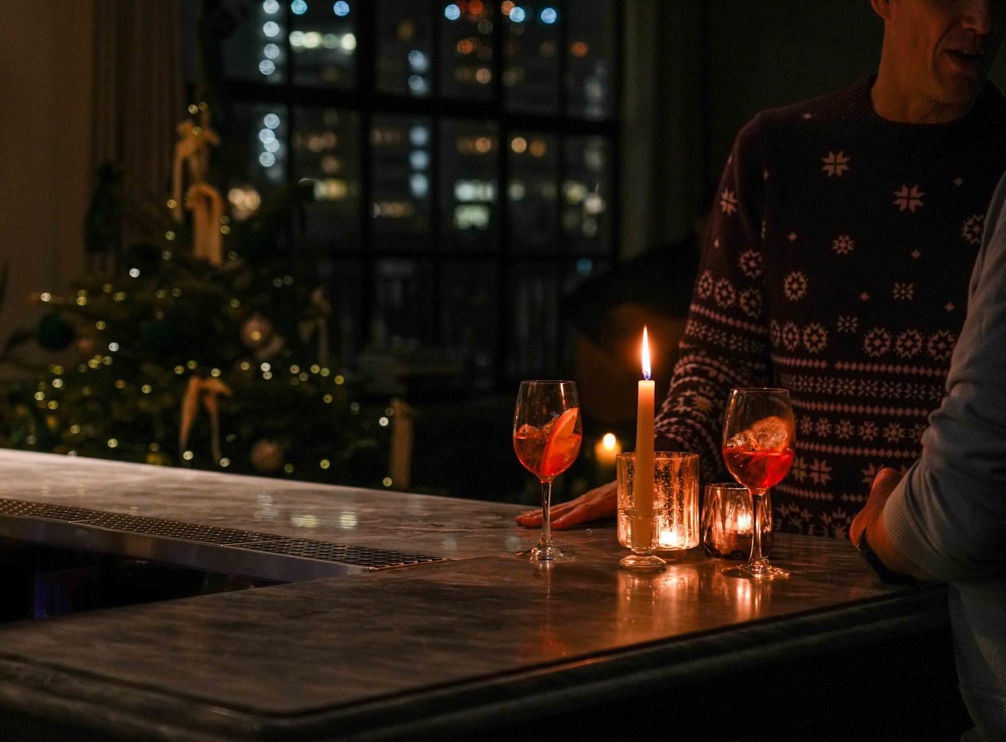 Two people enjoying festive cocktails by candlelight at Maslow’s rooftop bar in London, with a Christmas tree and city skyline in the background.