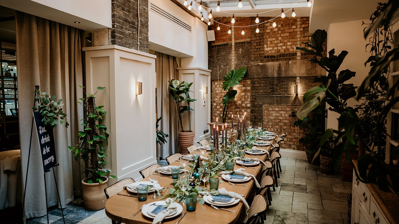 Elegant conservatory private dining space at Mortimer House in London, with long table, candlelight, hanging bulbs, and lush greenery.