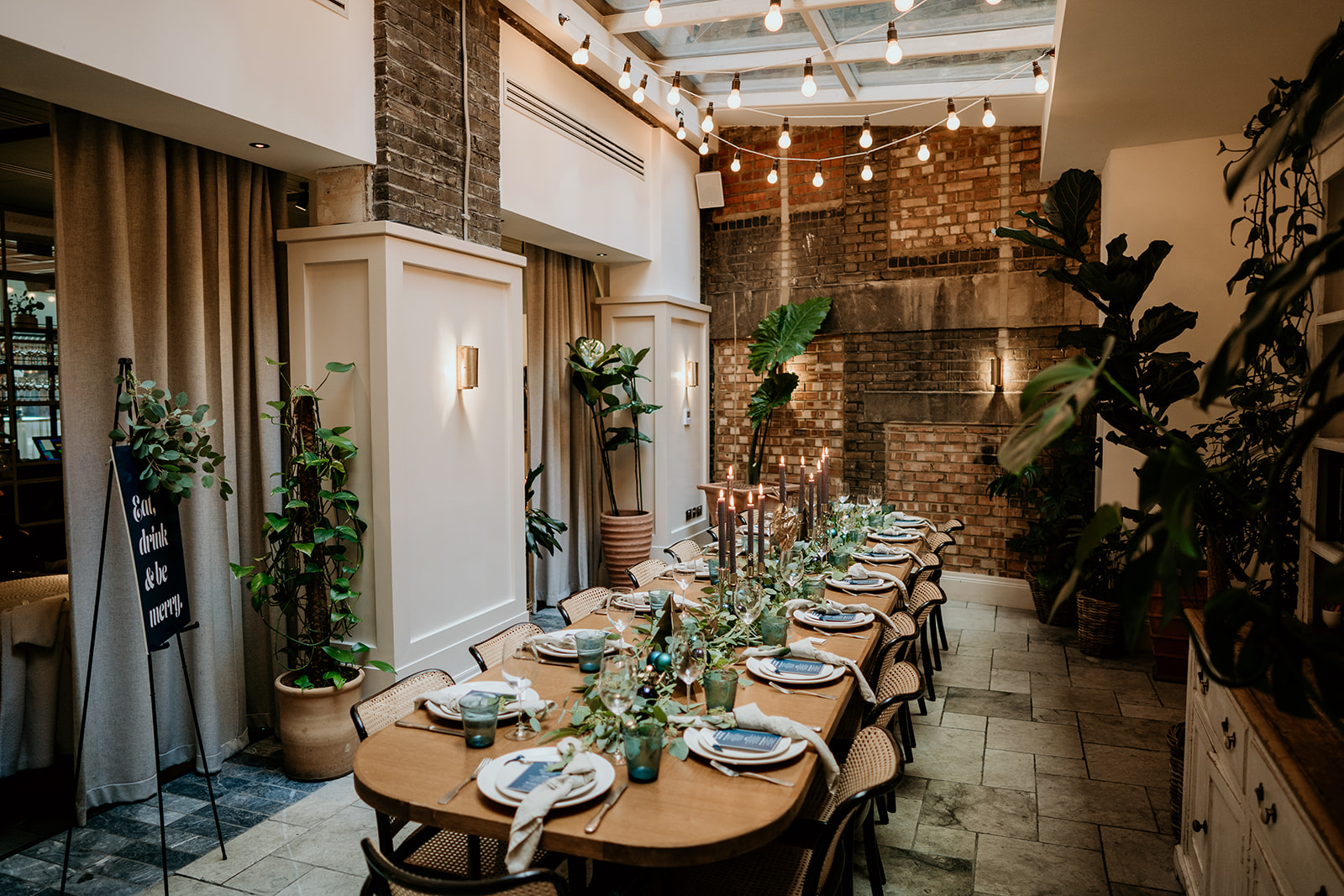 Elegant conservatory private dining space at Mortimer House in London, with long table, candlelight, hanging bulbs, and lush greenery.